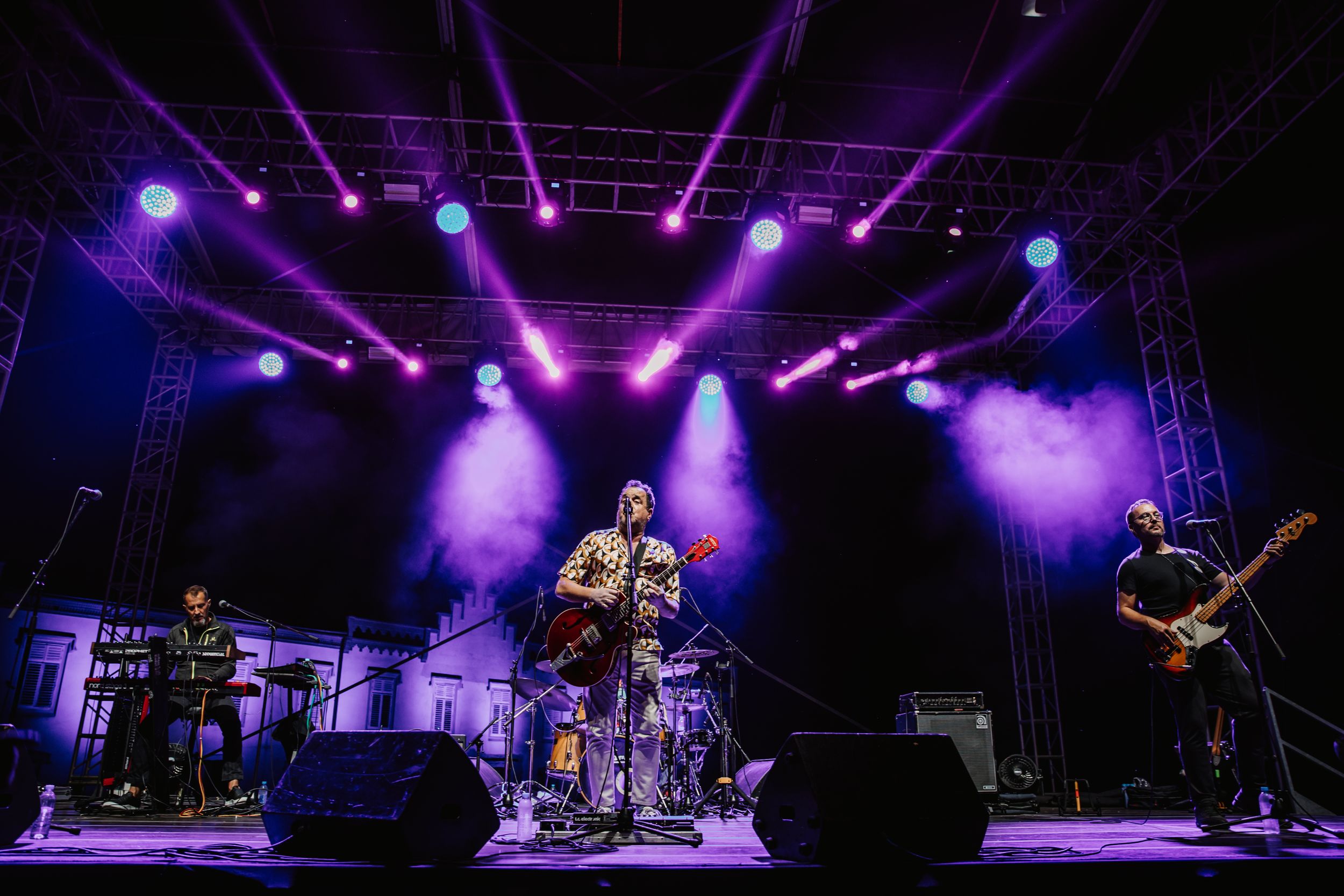 Wide shot of full band performing on concert stage with dramatic purple lighting and fog effects.