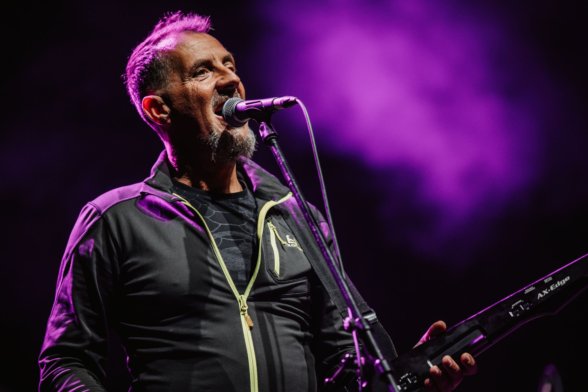 A performer sings passionately into a microphone against a purple-lit stage backdrop.