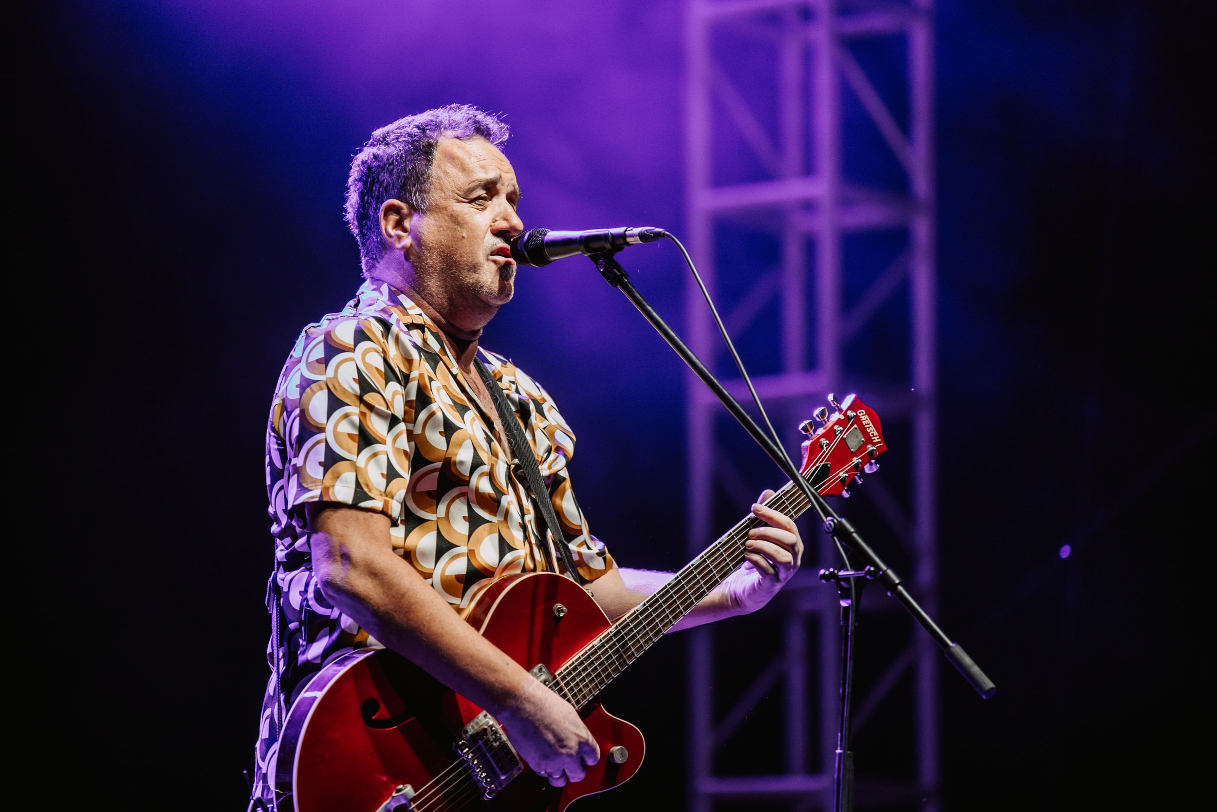 Musician performing on stage with a red guitar under dramatic purple lighting.