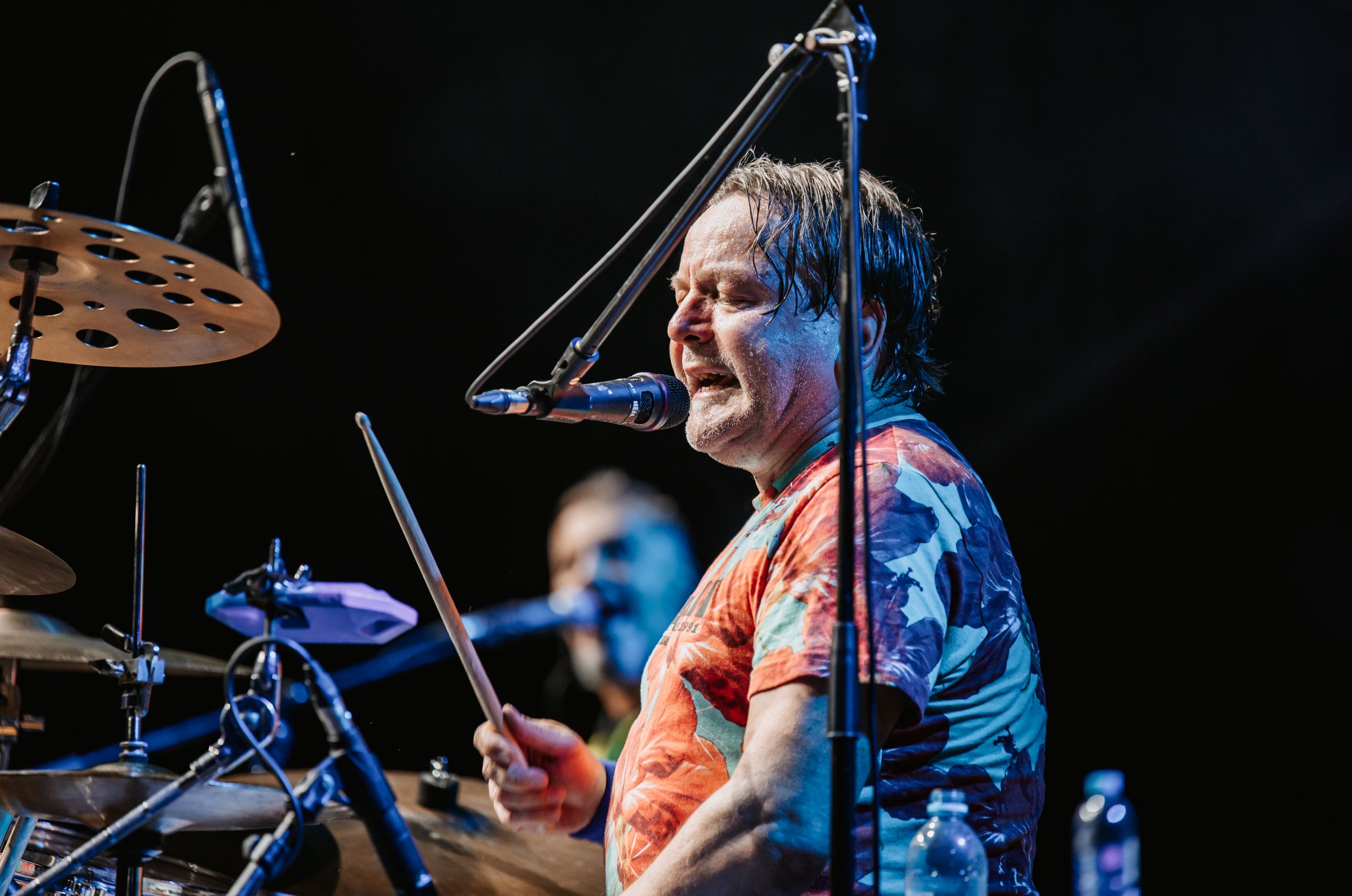 An energetic drummer in a tie-dye shirt performs behind a drum kit.