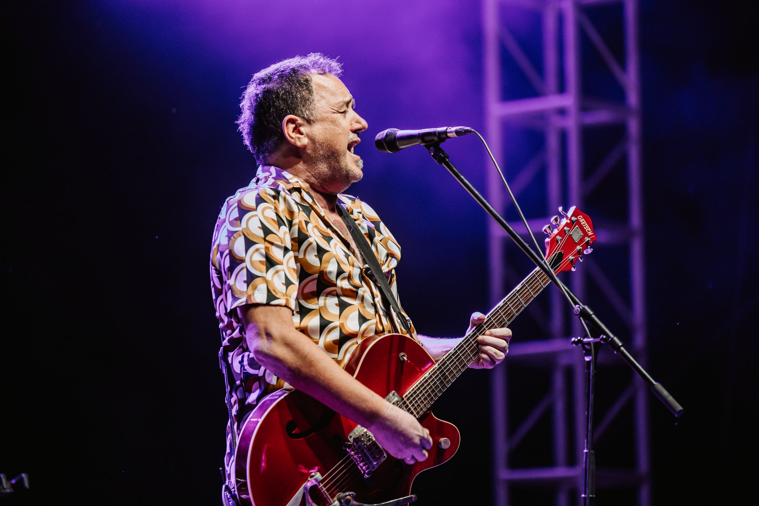 Musician performs energetically on stage with a red guitar under purple lighting during a concert performance.