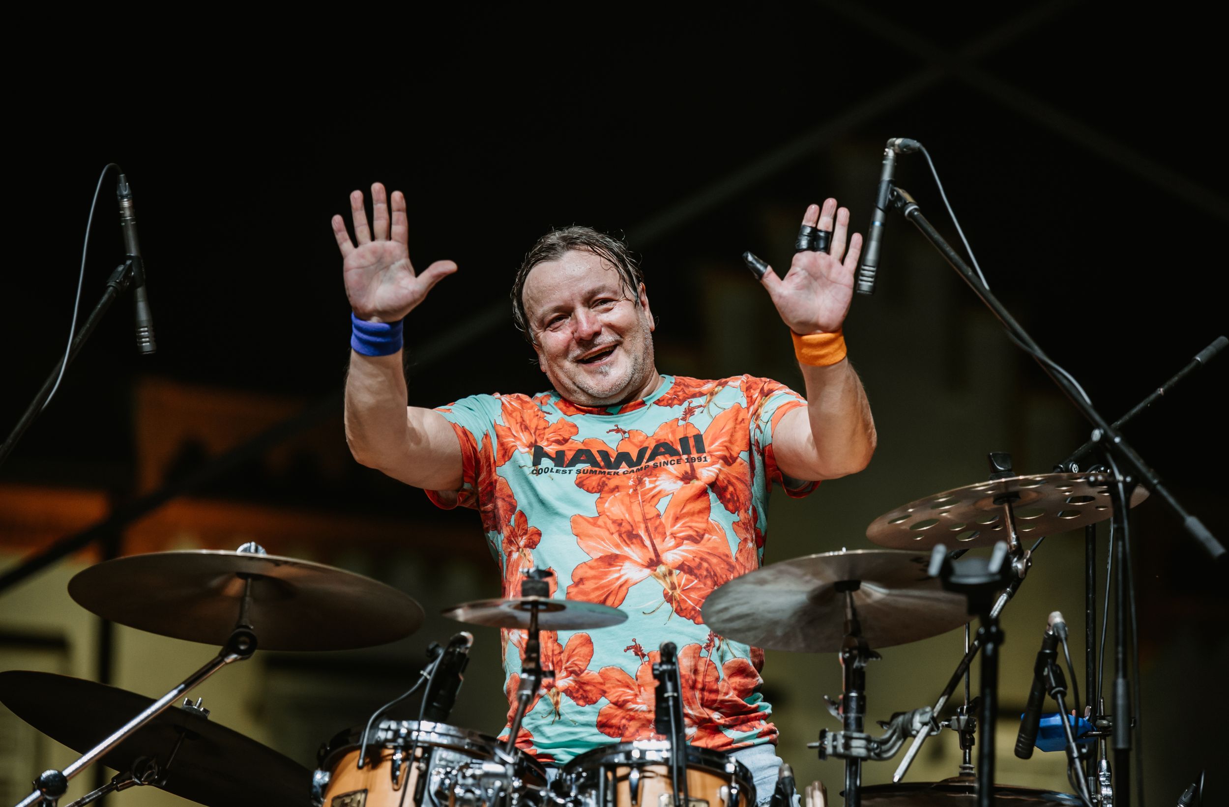 Drummer in vibrant floral shirt raises hands energetically behind drum kit during outdoor concert performance.
