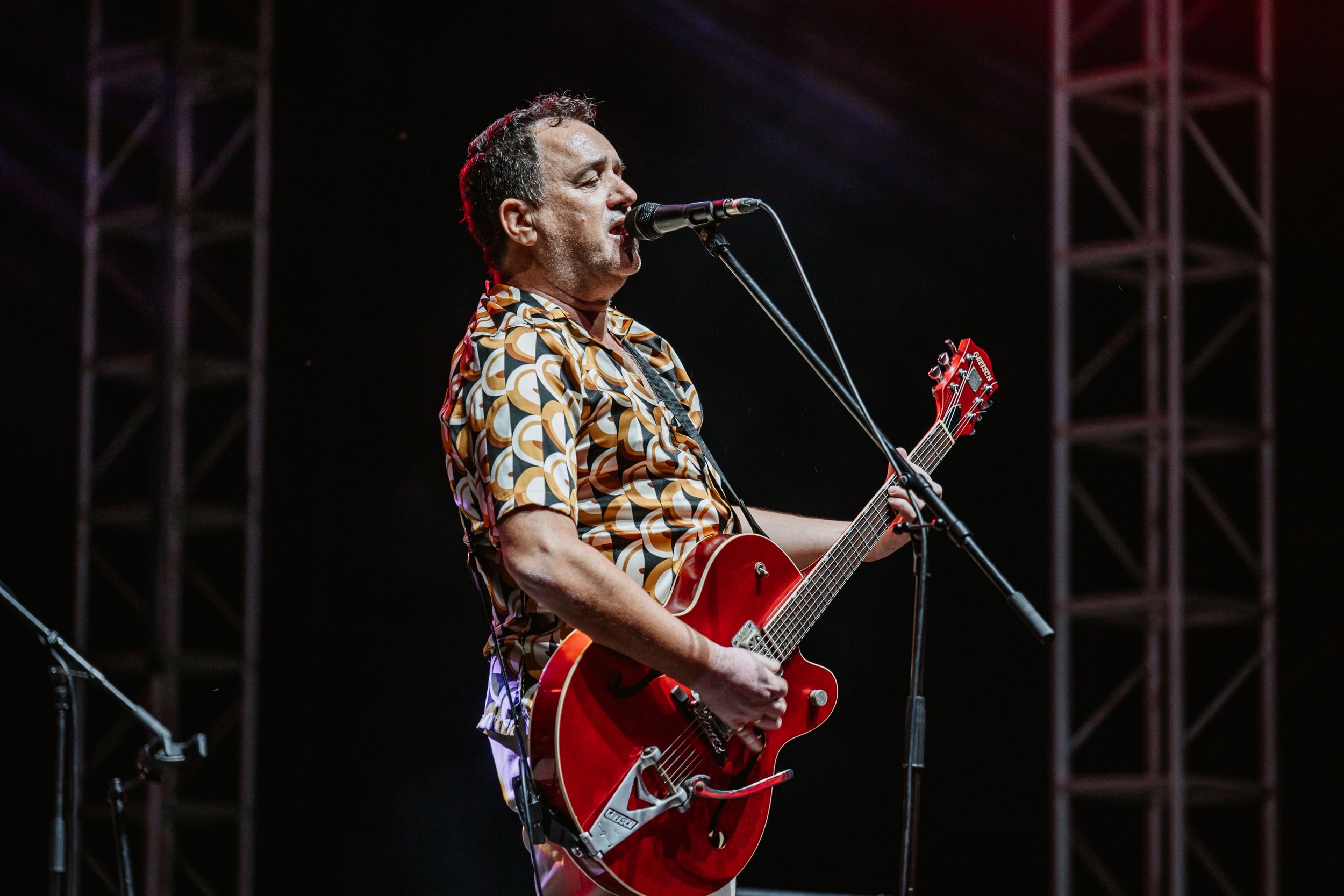 Musician in patterned shirt plays red electric guitar while singing into microphone on outdoor stage.