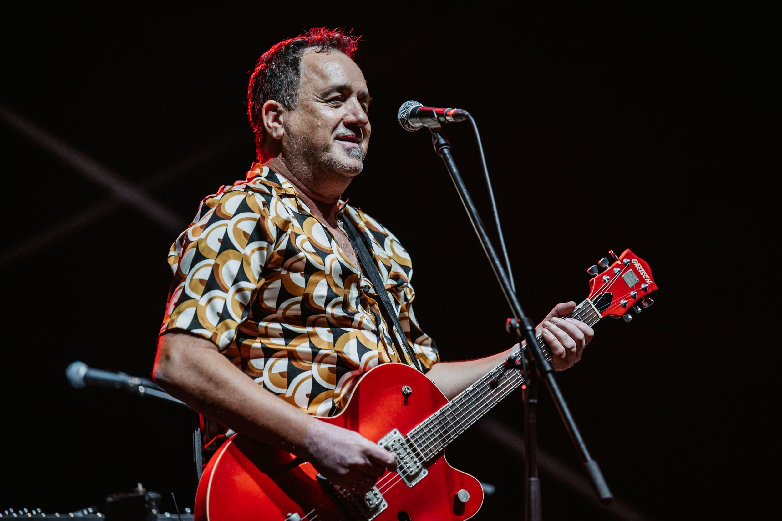 Guitarist performs with red electric guitar on illuminated stage wearing patterned short-sleeve shirt.