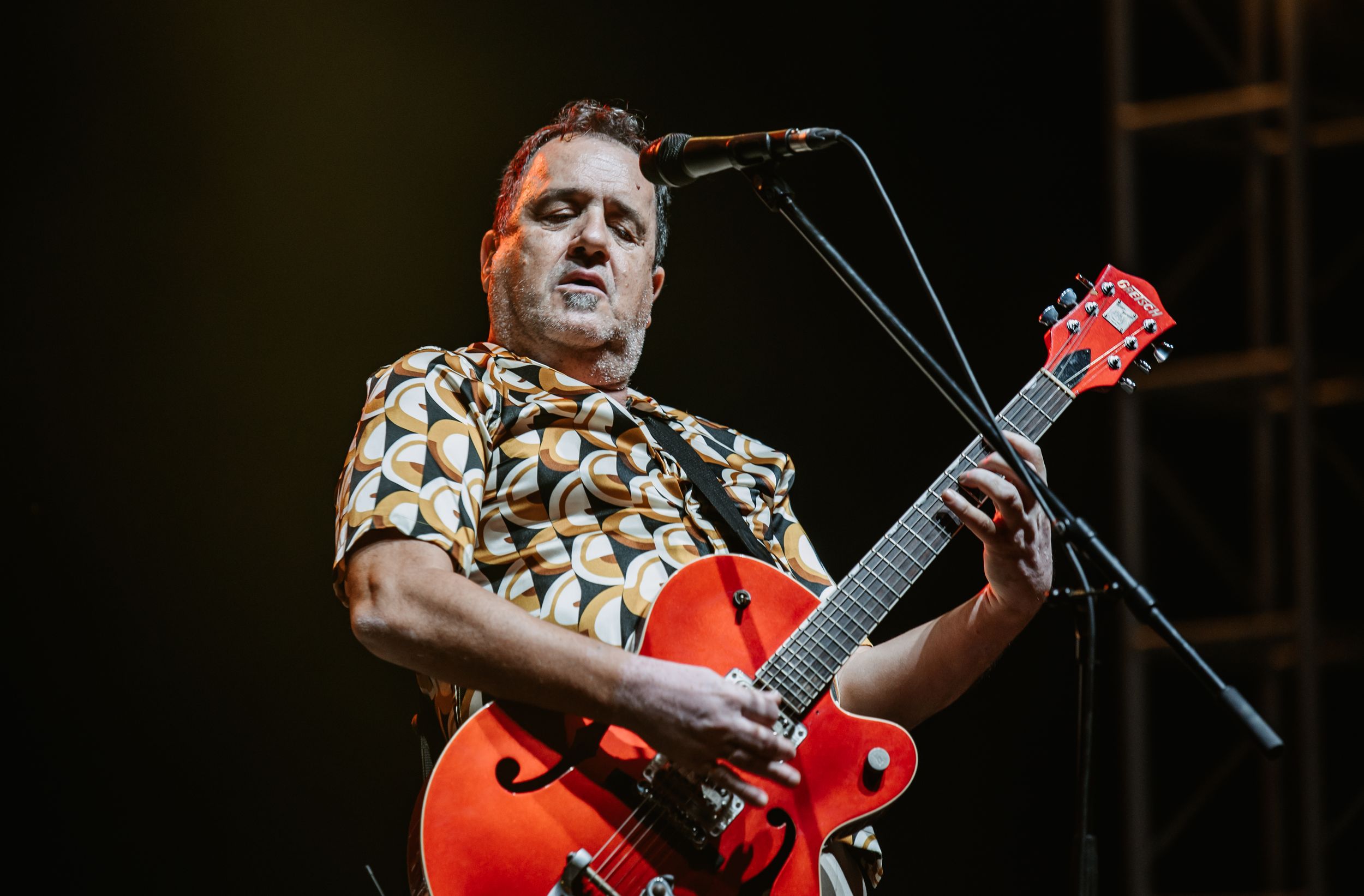 Performer plays bright red electric guitar while singing on dark concert stage.
