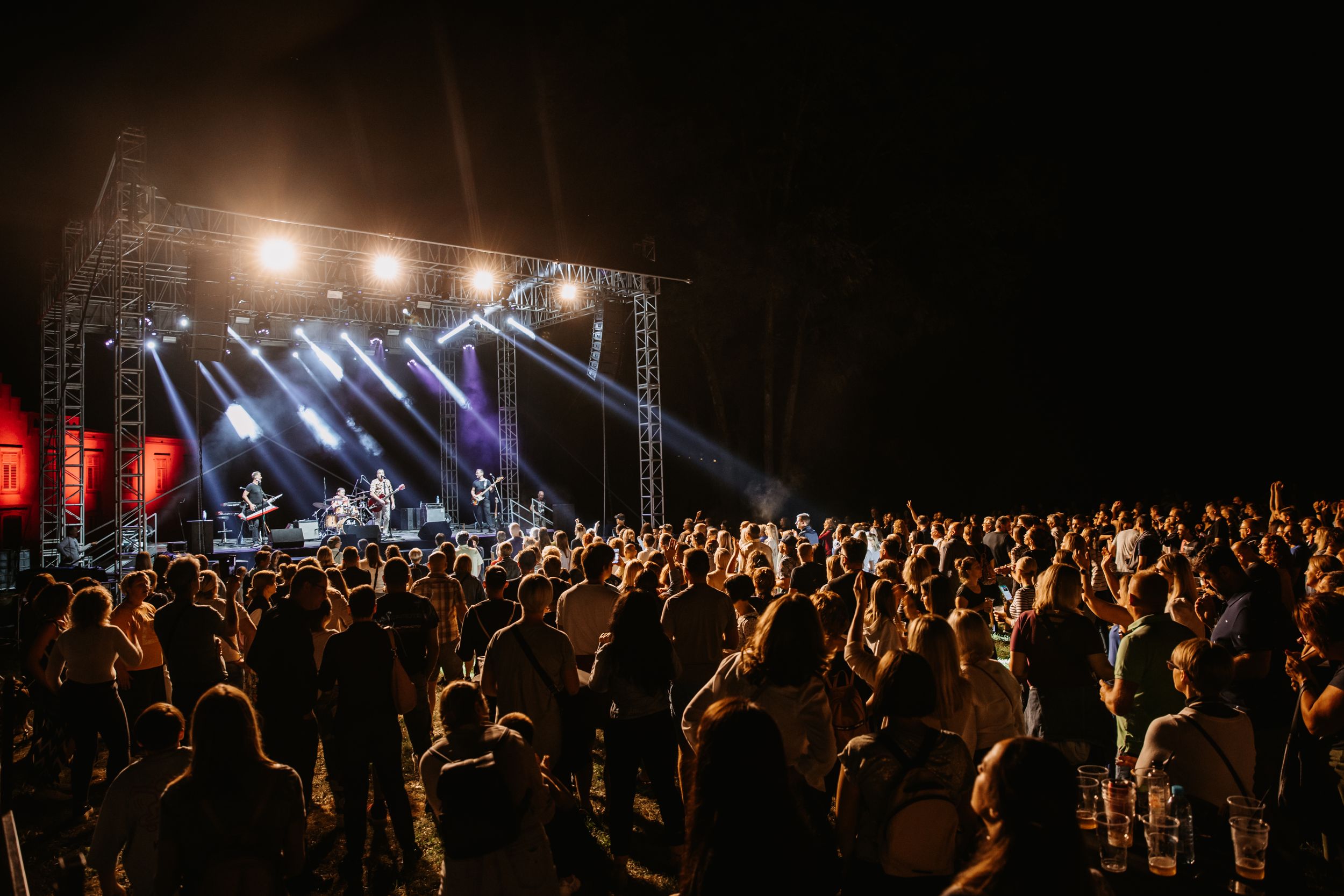 Large crowd at outdoor concert venue with dramatic stage lighting and metal truss structure.