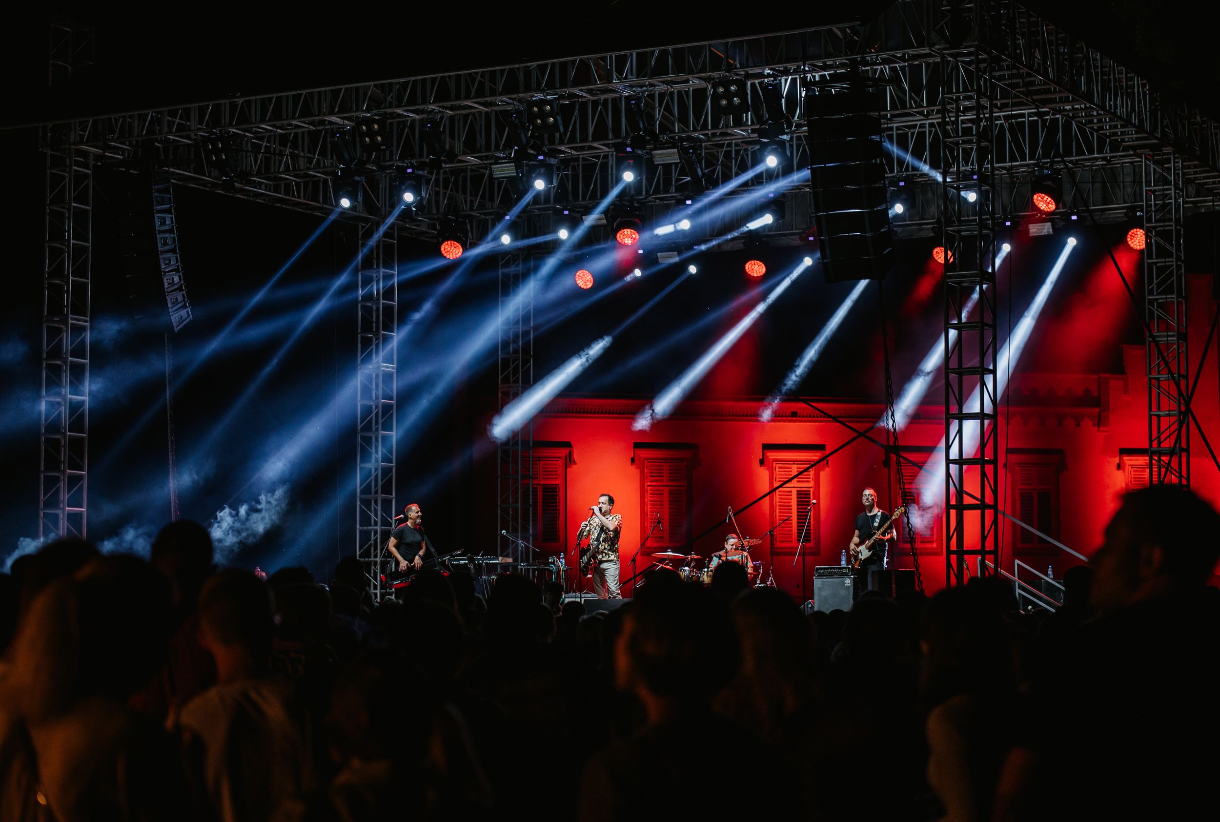 Concert stage with dramatic red and blue lighting beams cutting through smoke against dark sky.