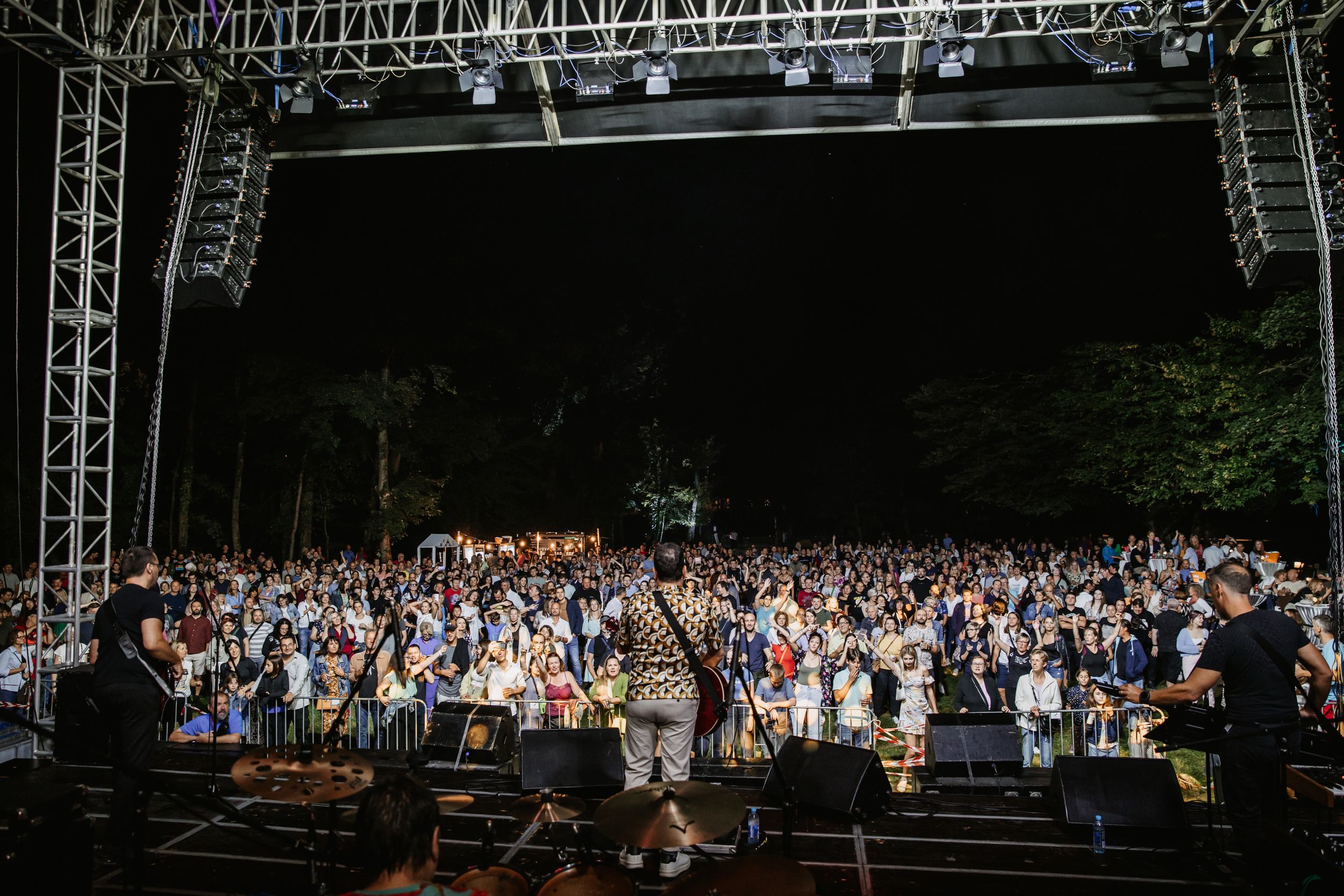 Wide shot of outdoor concert showing large crowd and stage setup with metal truss rigging.