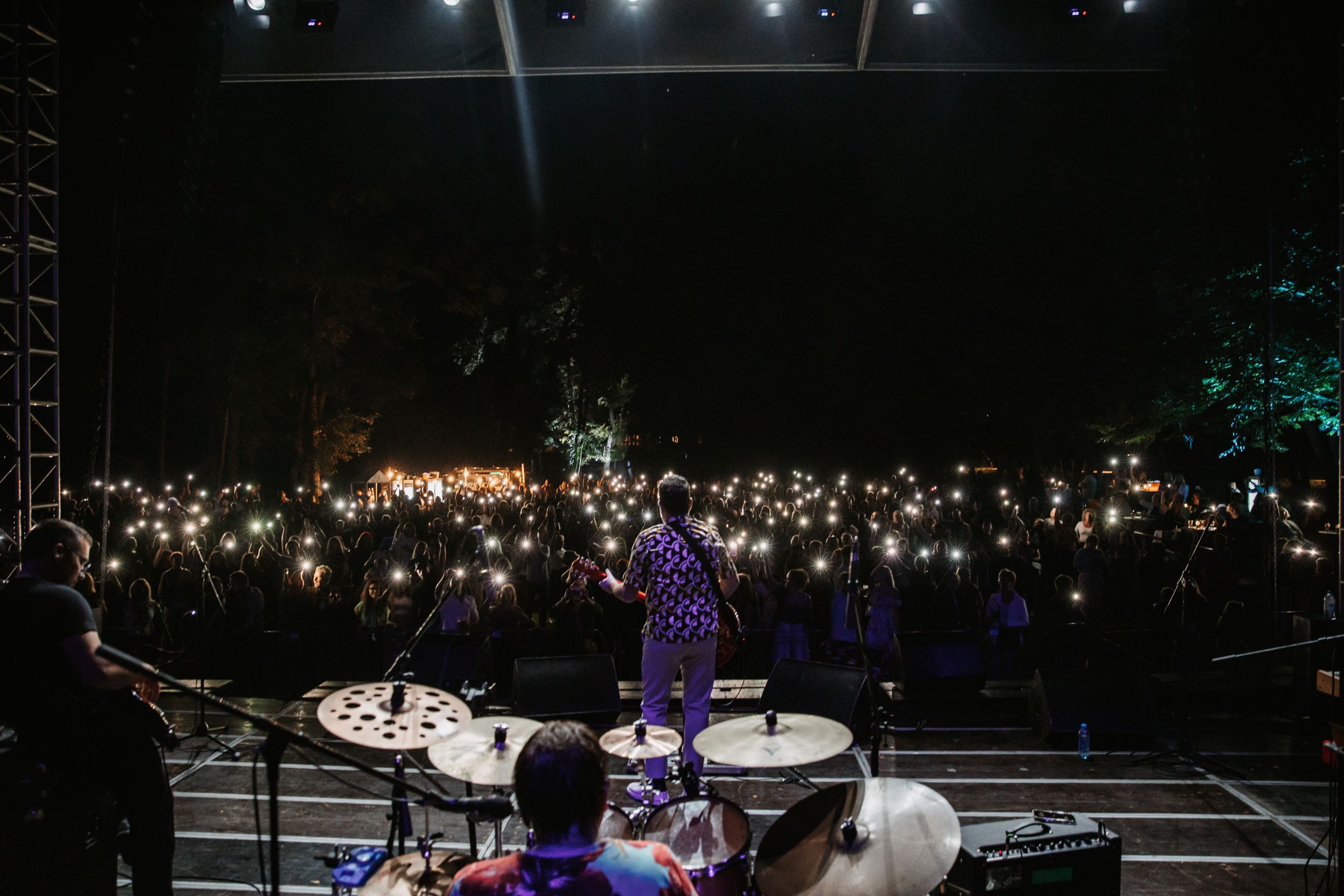 Concert performer on stage viewed from behind with crowd holding up phone lights in darkness.