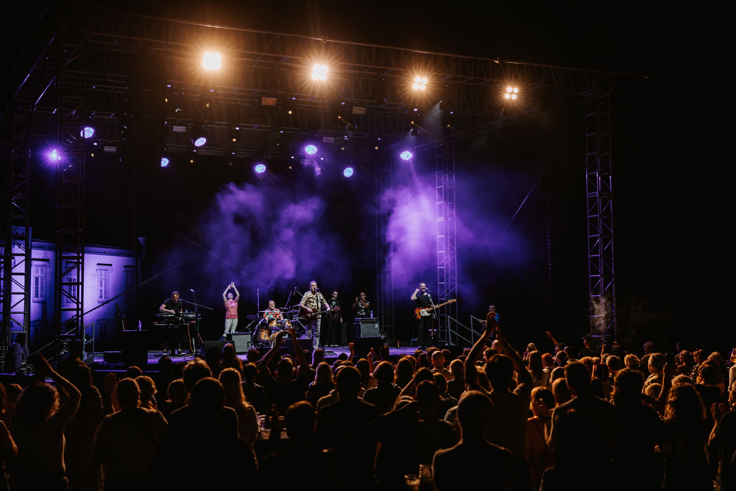Band performing on purple-lit stage with silhouetted crowd in foreground.