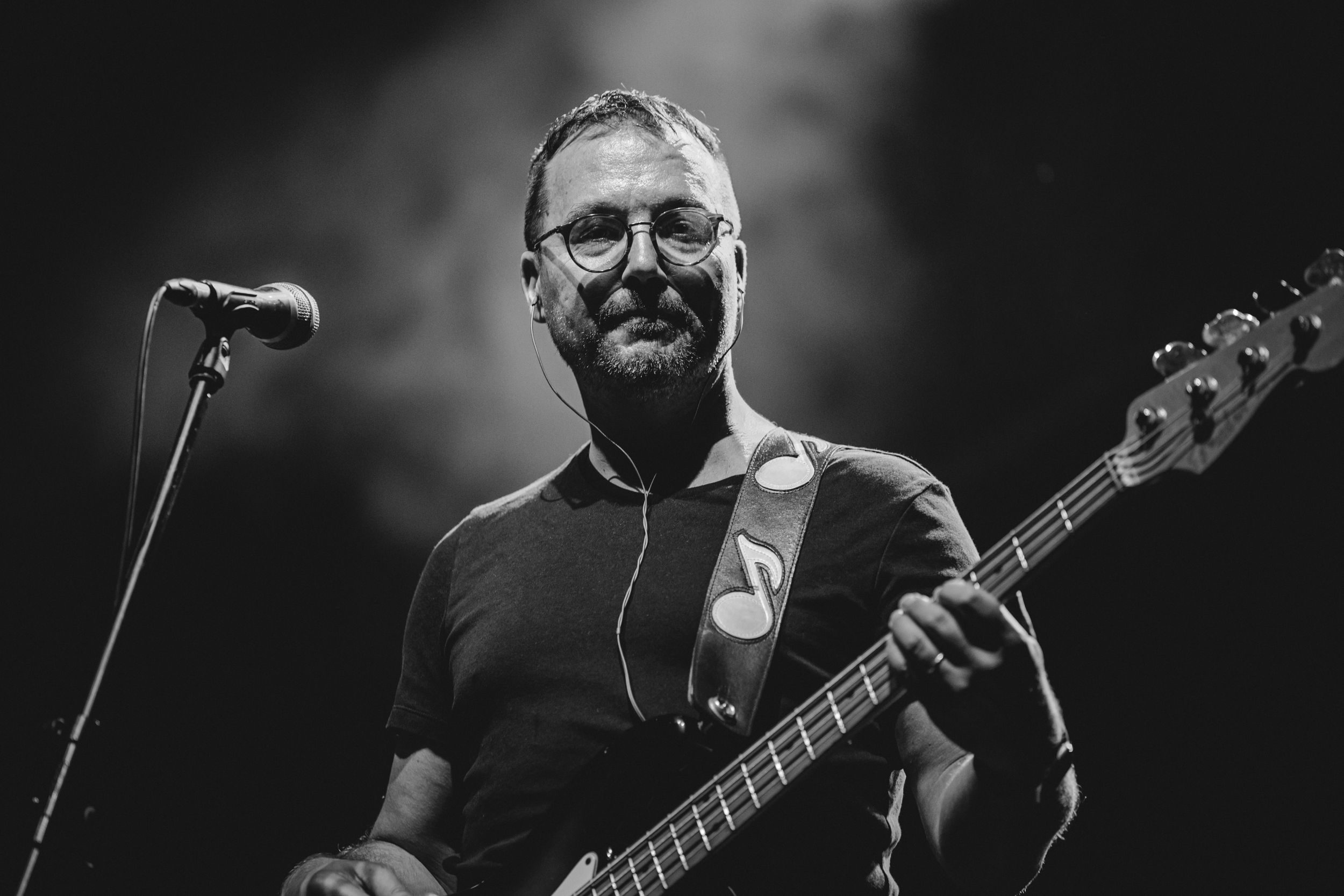 Bass guitarist performs in black attire on stage in dramatic black and white lighting.