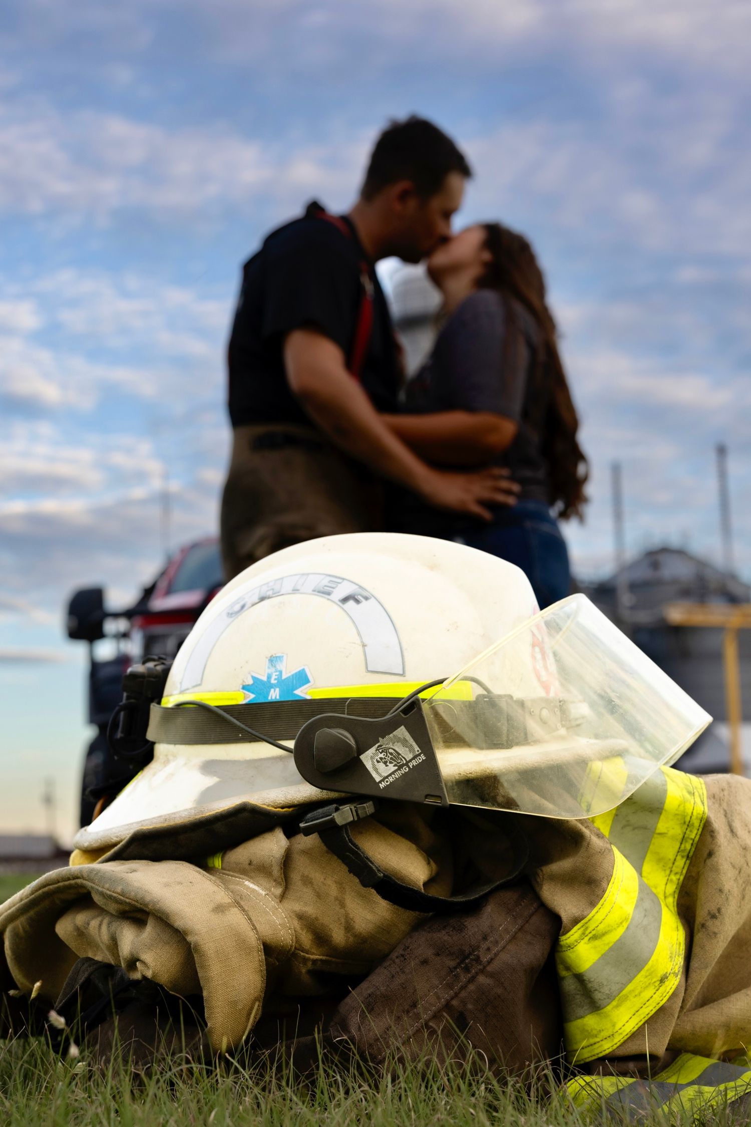 Firefighter helmets rest on grass while a couple shares an intimate moment in the background.