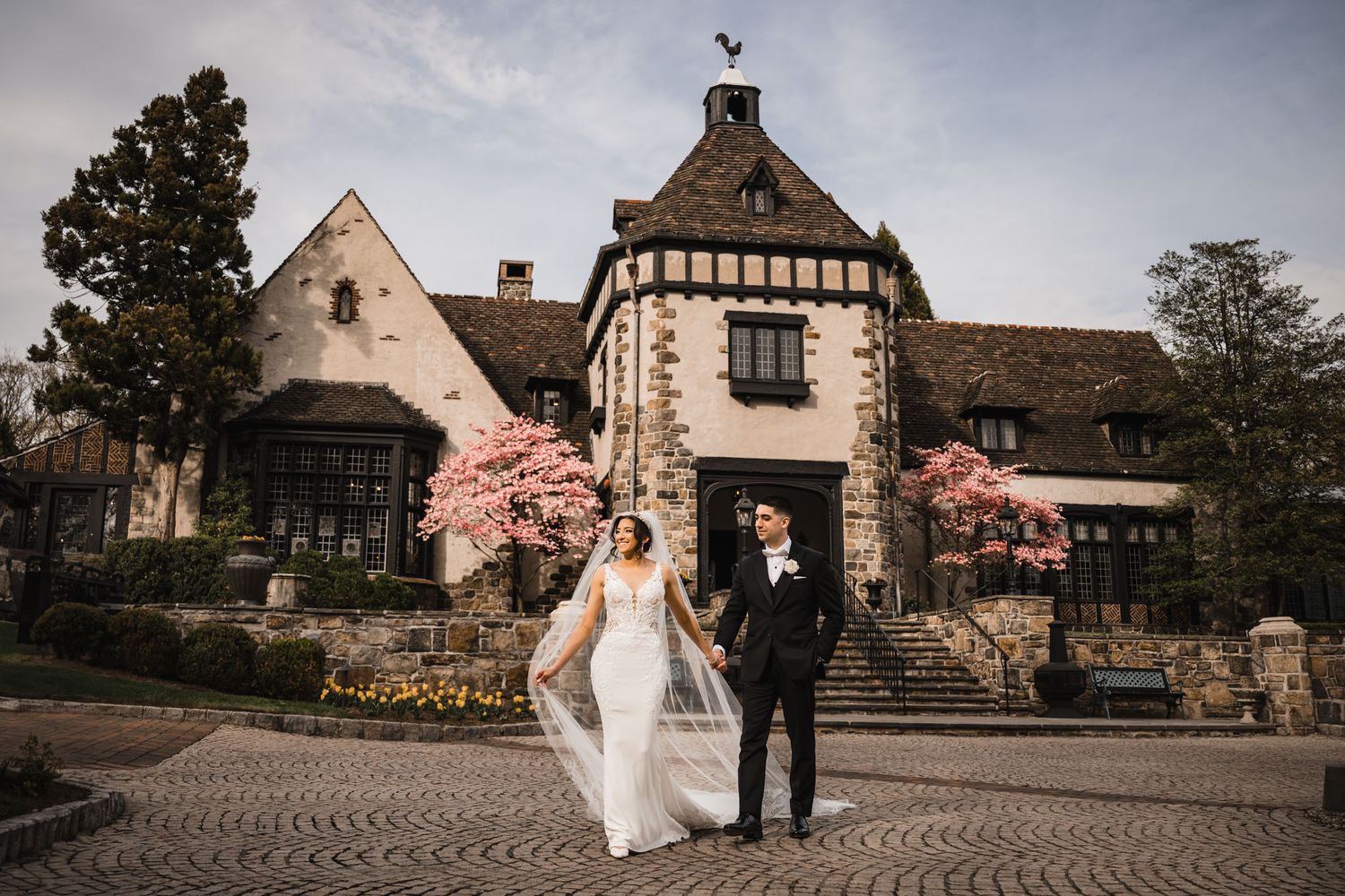 Elegant couple stands before a historic Tudor-style mansion with blooming cherry trees and stone facade.