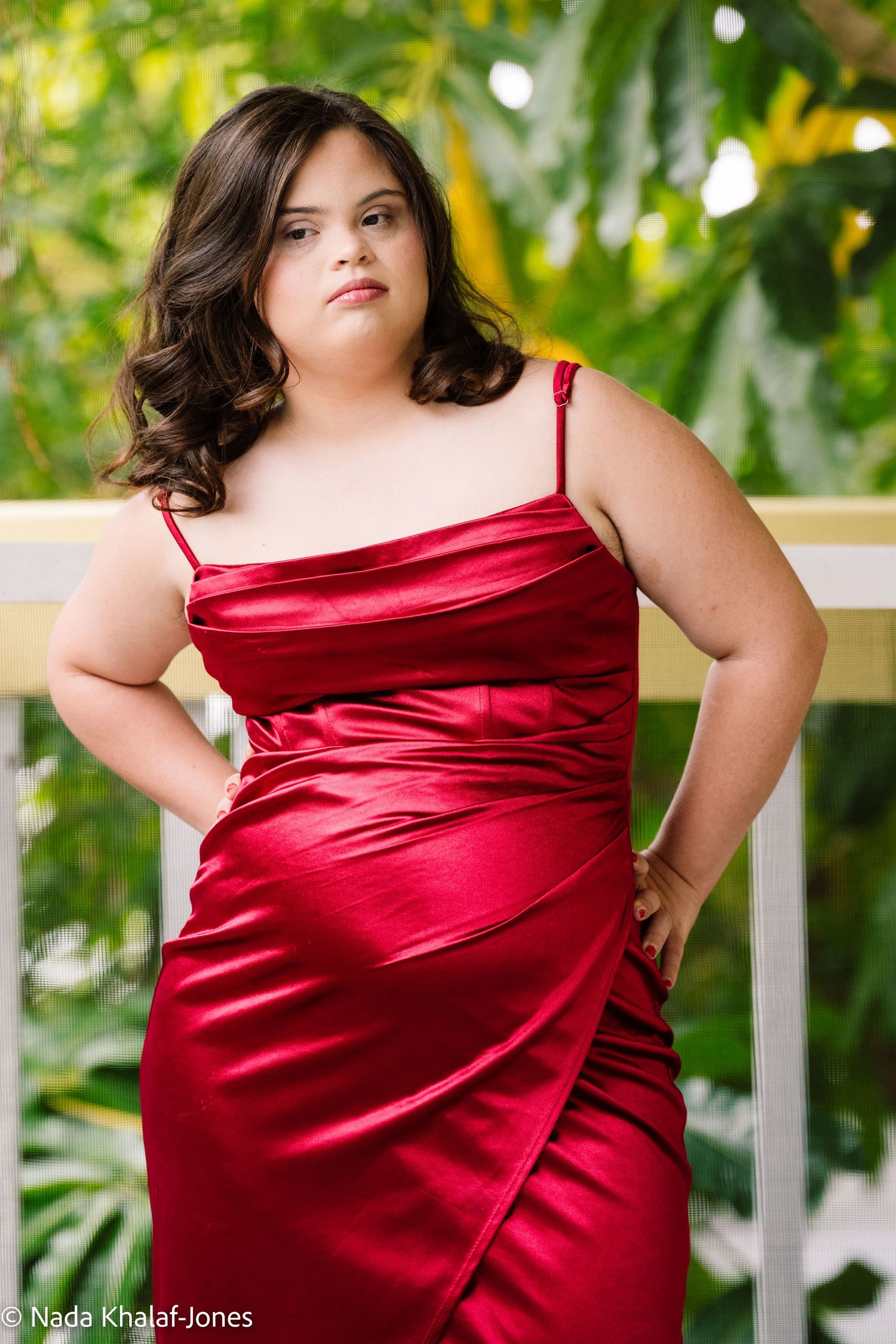 A person in a flowing red satin gown poses against a white railing with lush greenery in the background.