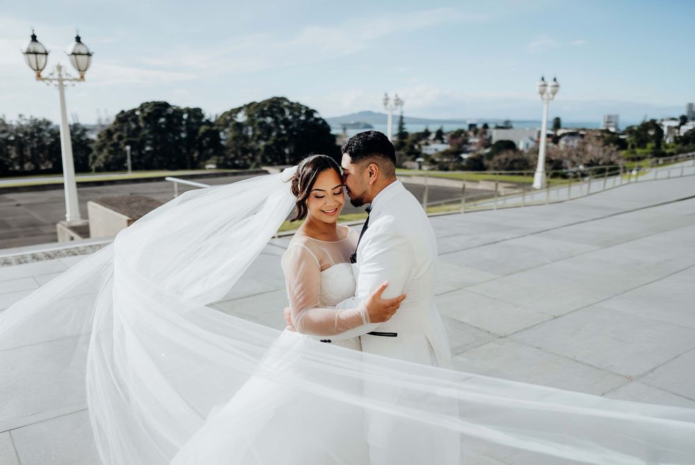 A bride and groom embrace on white steps, her veil flowing dramatically in the wind against a scenic waterfront backdrop.