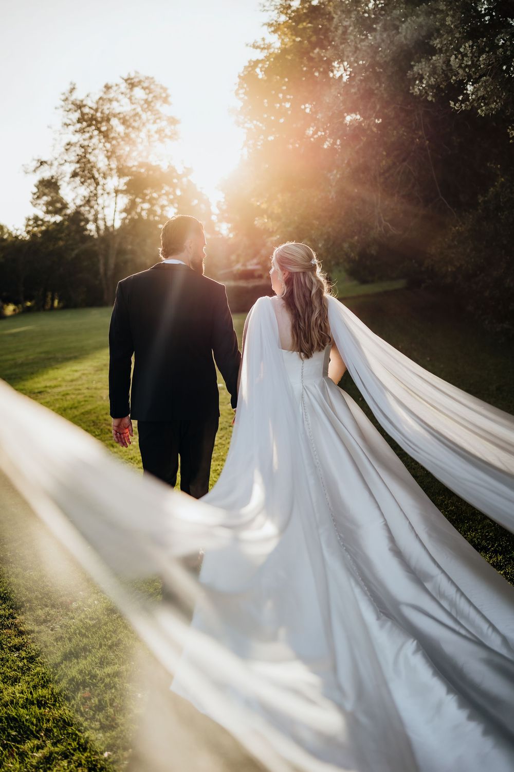 A wedding couple walks through a sunlit garden as the bride's long white veil flows dramatically in the breeze.