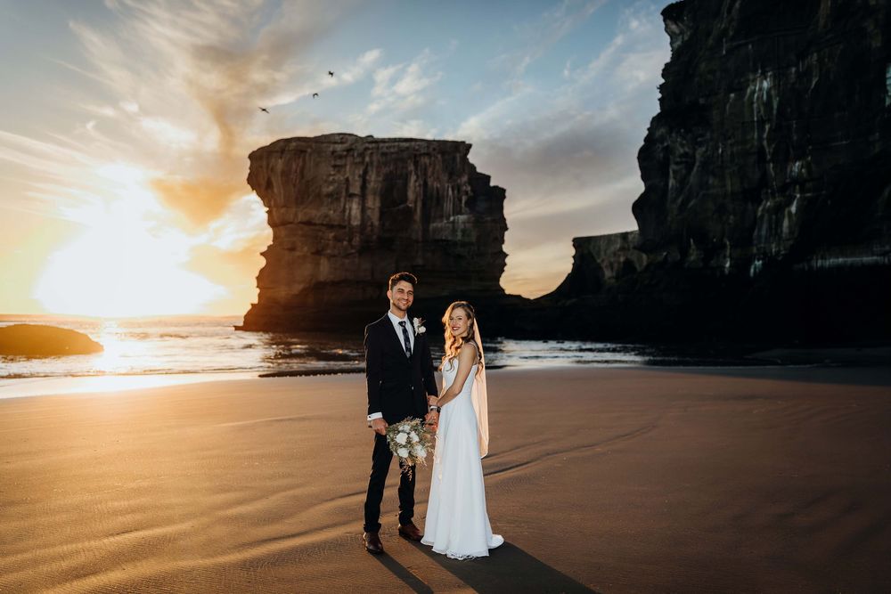 Couple holds hands on sandy beach at sunset with dramatic rock formations behind them in a romantic wedding photo.