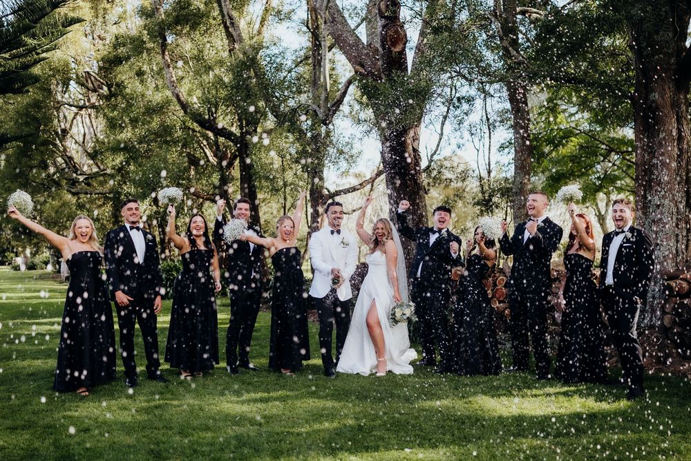 Wedding party celebrating outdoors with confetti, bride and groom in white surrounded by guests in black formal attire under trees.