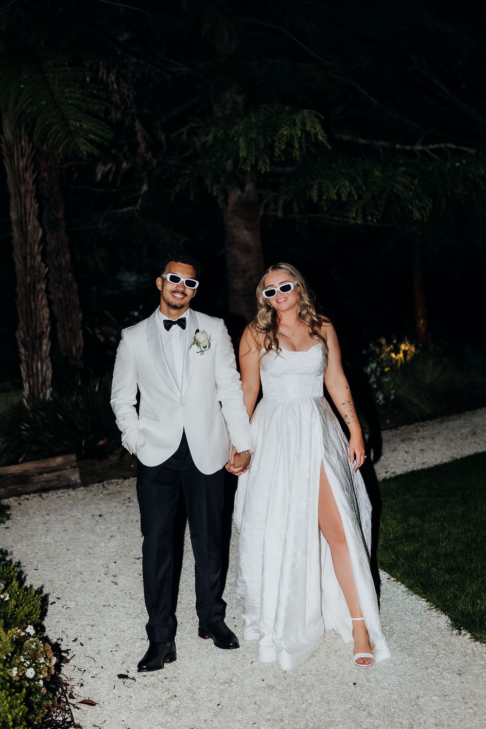 Wedding couple in white sunglasses pose at night, groom in white tuxedo and bride in elegant slit gown.