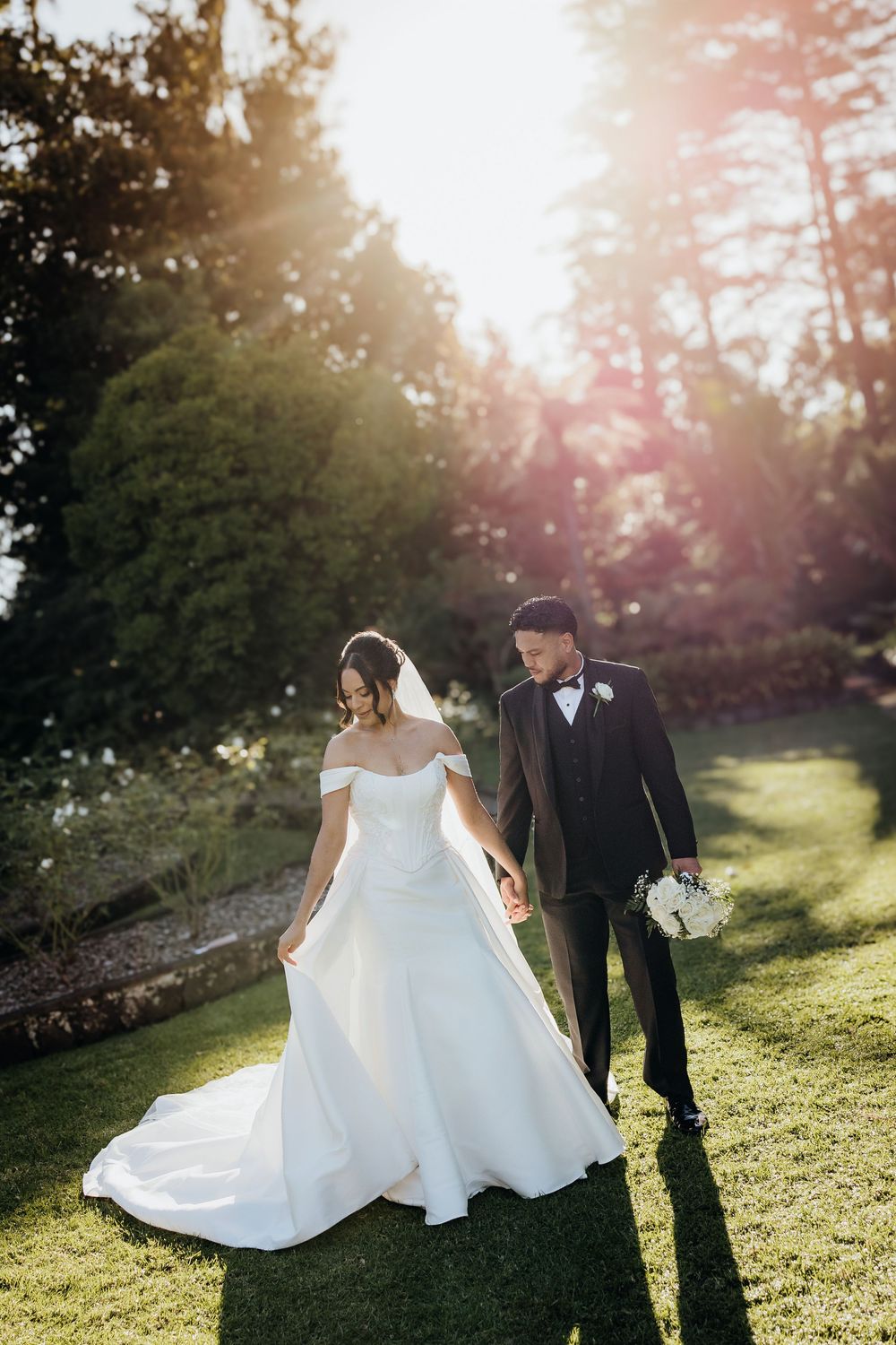 Bride and groom holding hands walking on green lawn with golden sunlight filtering through trees.