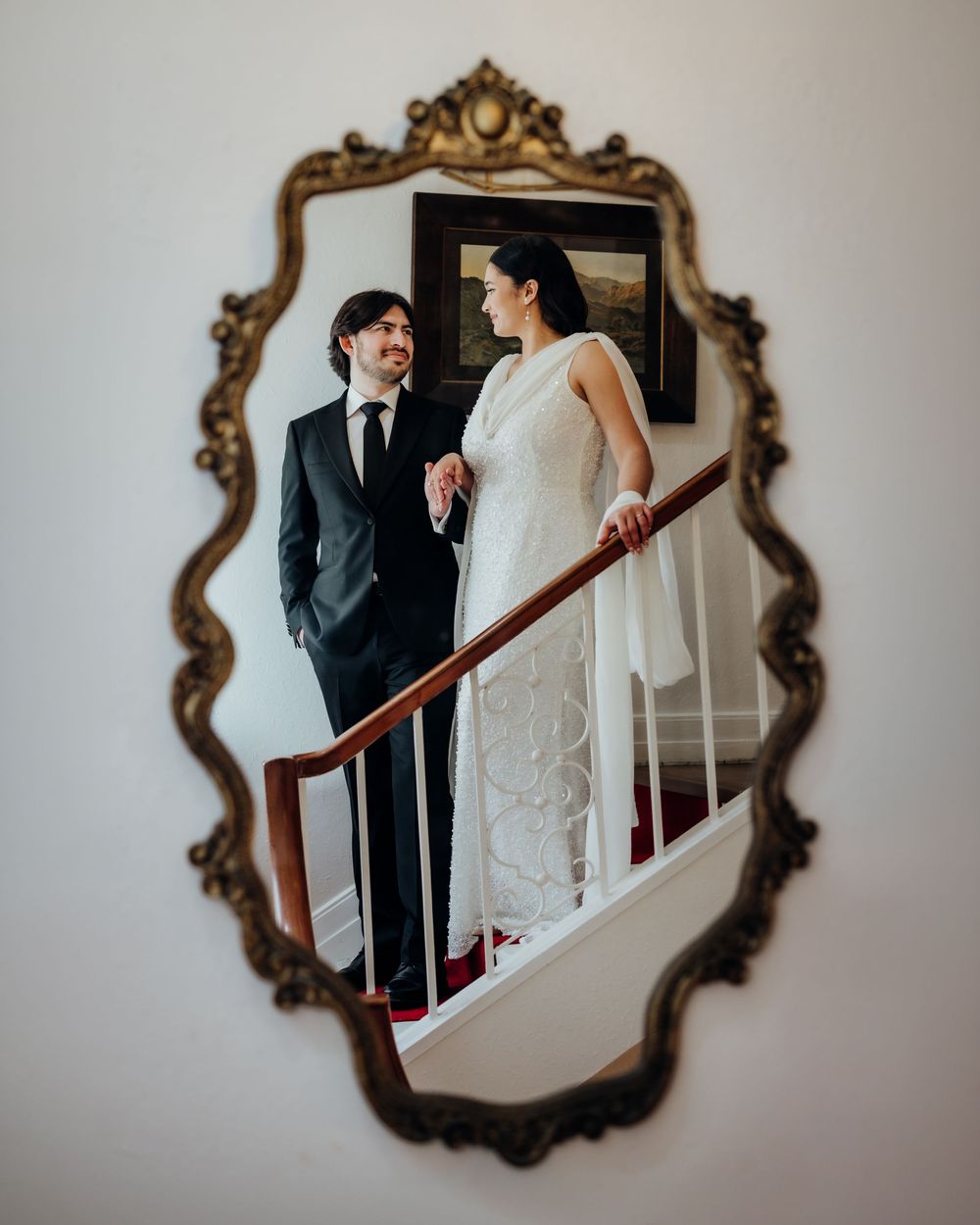 Bride and groom reflected in ornate gold mirror on staircase, bride in white gown, groom in dark suit.