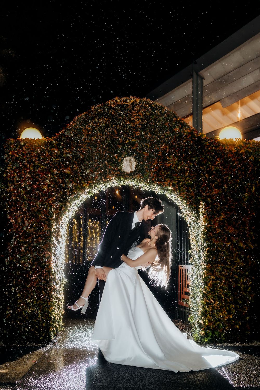 Bride and groom share a romantic dip kiss under a glowing floral arch at night with sparkling lights.