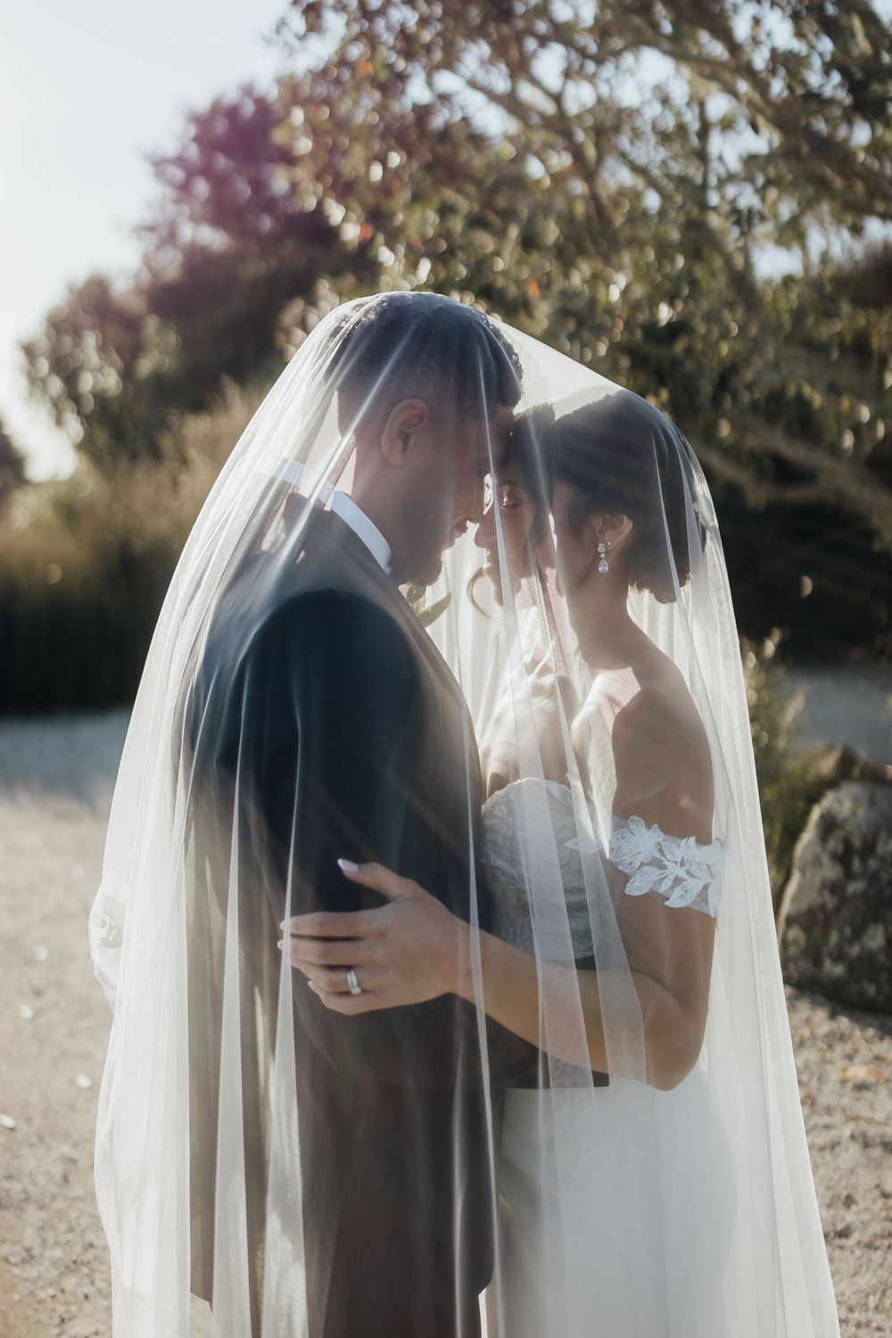 Bride and groom share an intimate moment under a flowing white veil outdoors on their wedding day.