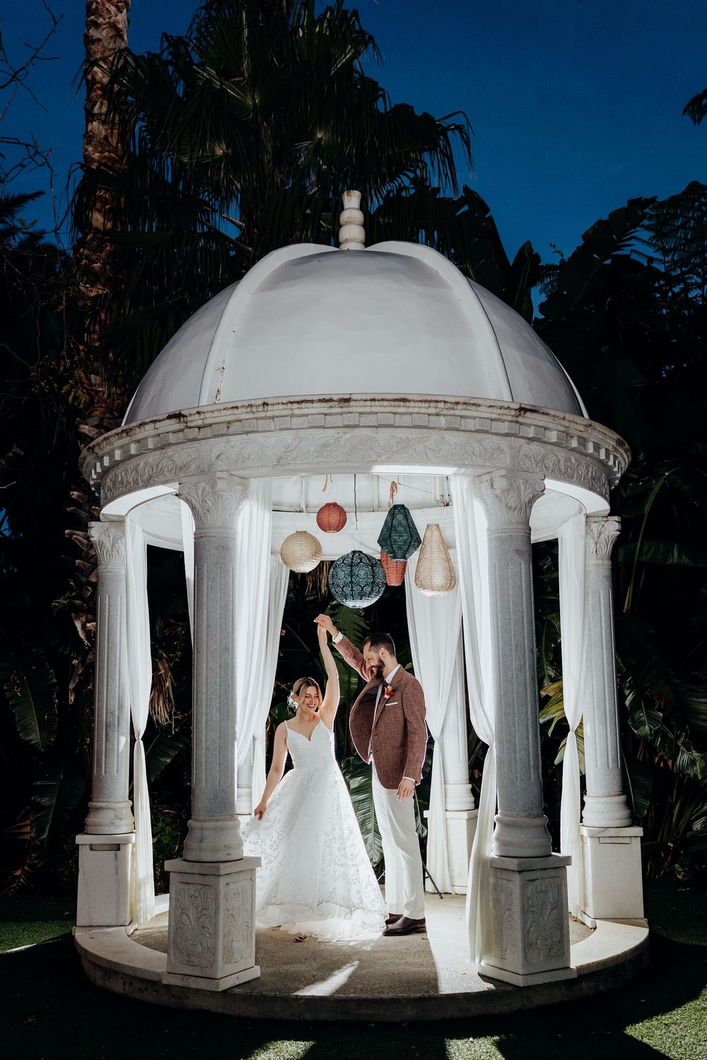 Bride and groom dancing under an elegant white gazebo adorned with colorful lanterns at night.