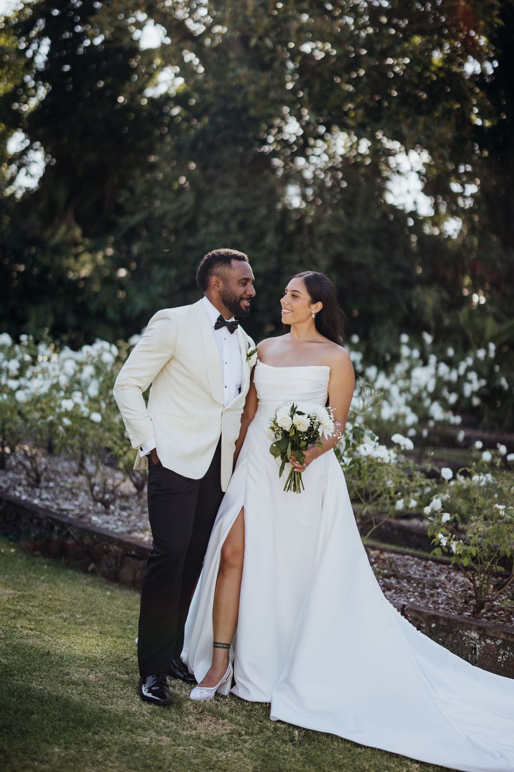 A romantic wedding couple sits together on grass, sharing intimate moments in their elegant white attire.