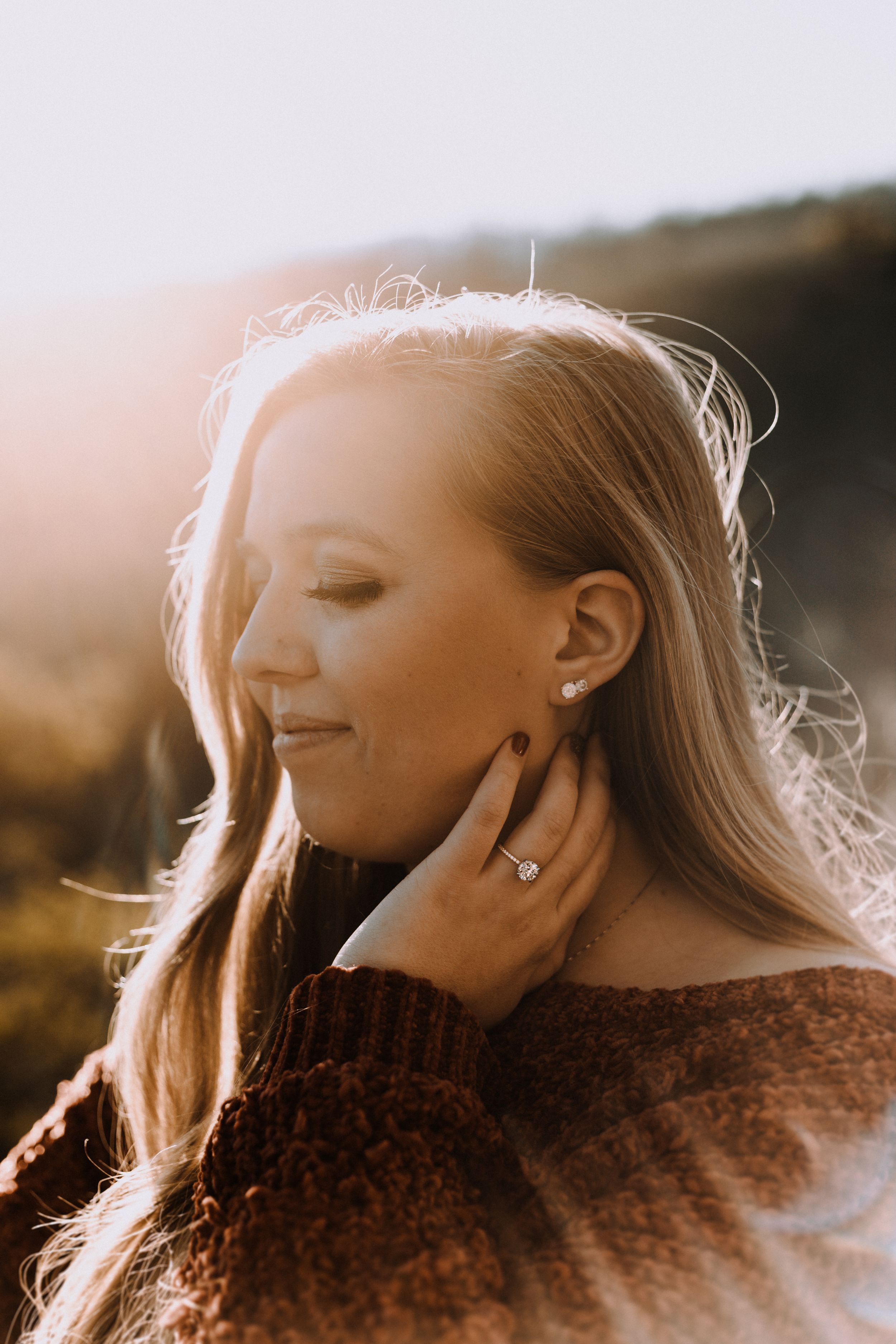 Woman with long blonde hair in brown sweater poses serenely in golden sunset light.