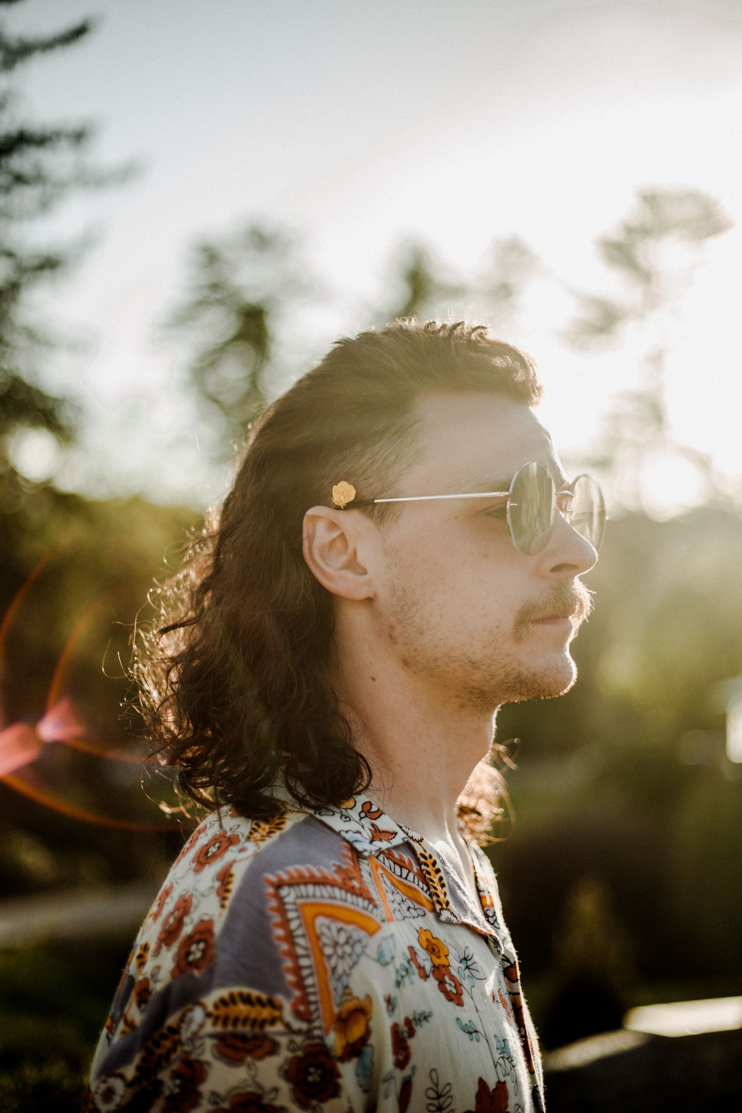 Person with curly hair and sunglasses in patterned shirt silhouetted against sunset through trees.