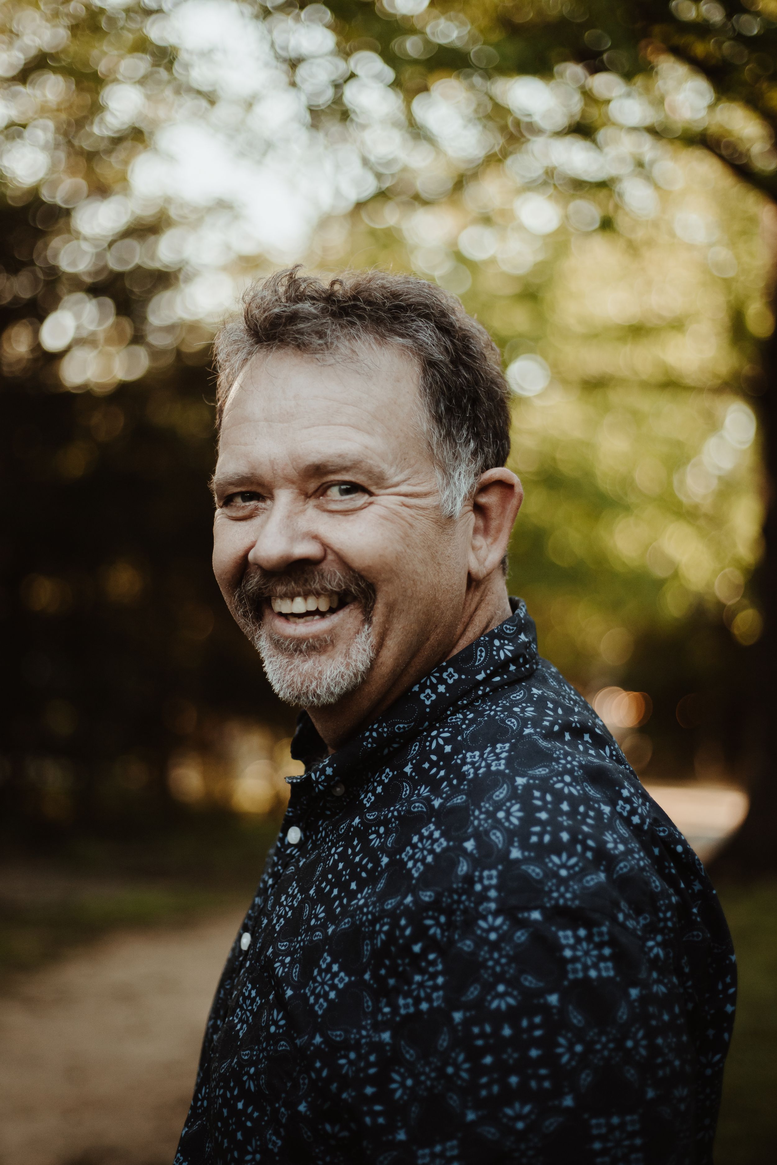 Candid portrait of person in patterned shirt smiling outdoors with bokeh background.