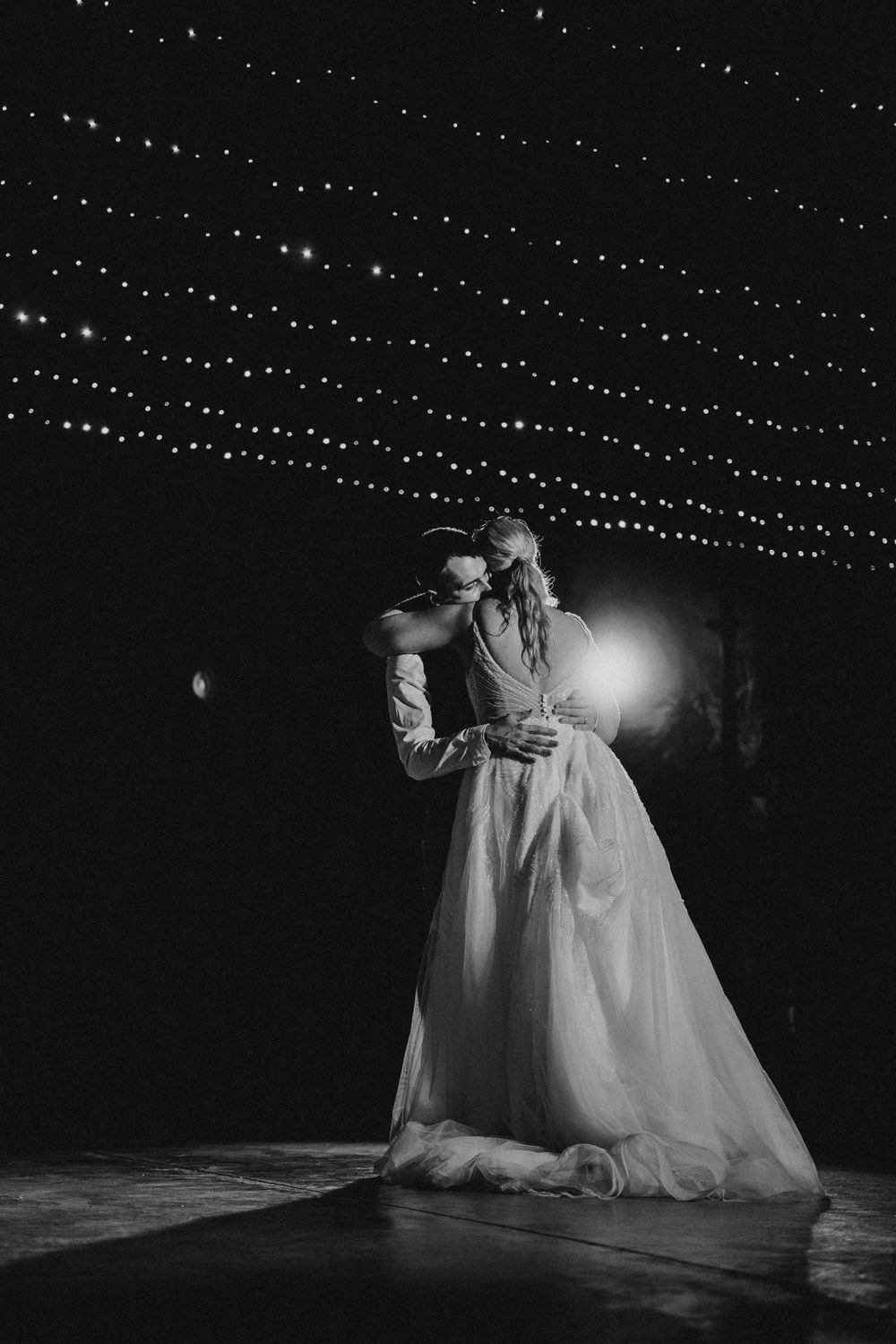 A romantic first dance at a wedding reception under twinkling string lights in black and white photography.