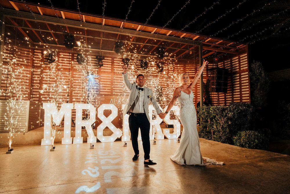 A couple celebrates their wedding reception in front of illuminated MR & MRS letters under a wooden pergola at night.