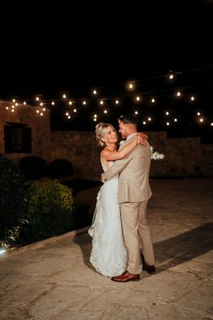 A couple shares their first dance under romantic string lights at an outdoor stone venue during their evening wedding celebration.