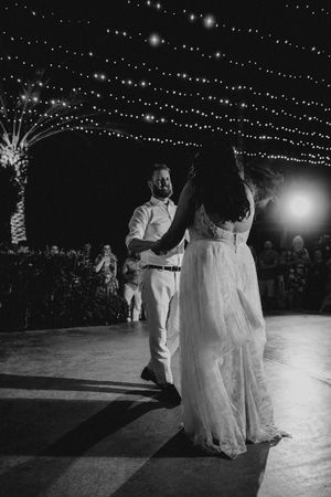 A couple shares their first dance at an outdoor wedding reception under twinkling string lights in black and white.