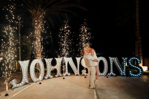 A sparkler-lit wedding display with illuminated JOHNSONS text sign and a couple posing in front at night.