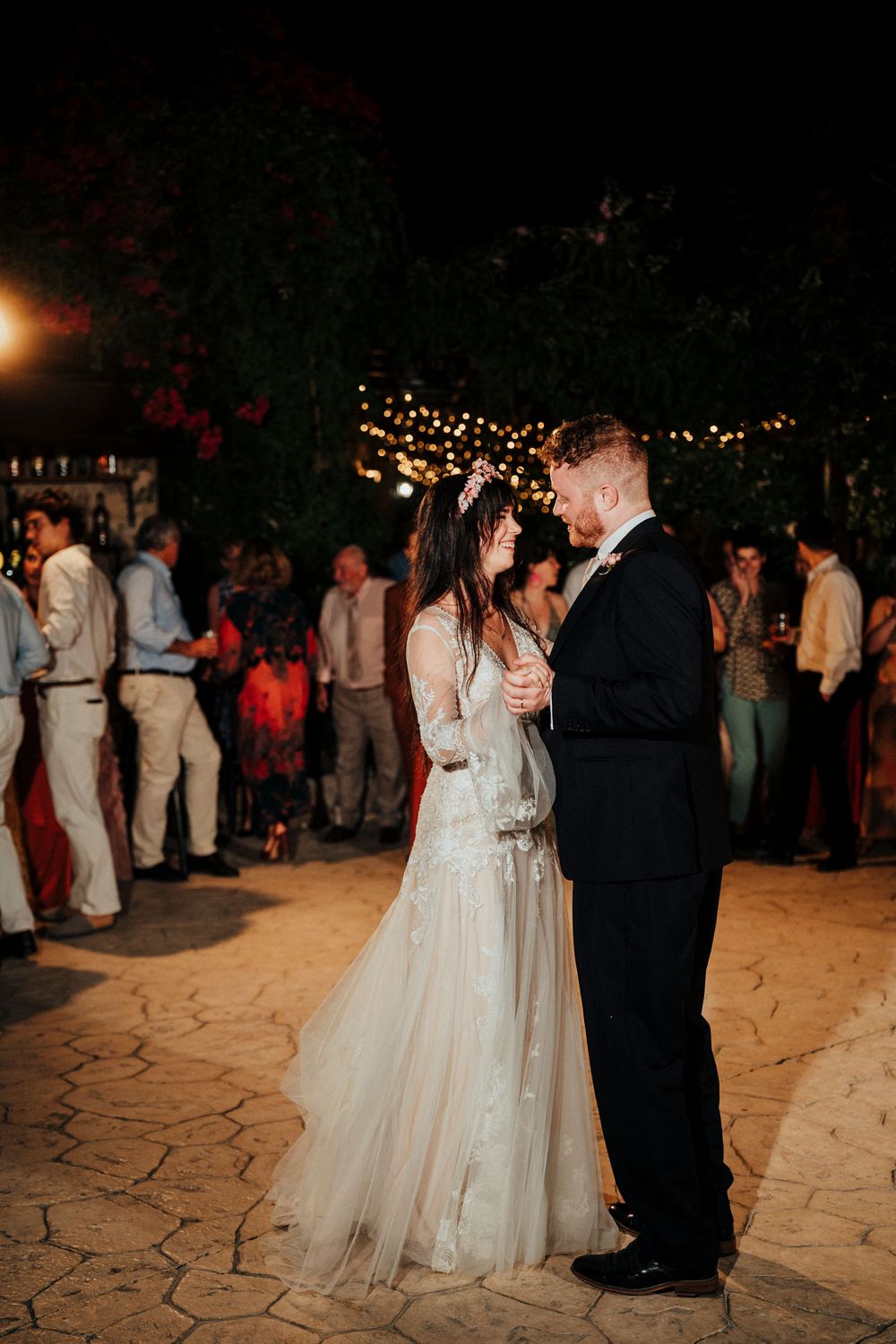 A couple shares their first dance at an outdoor evening wedding reception under twinkling lights on a stone patio.