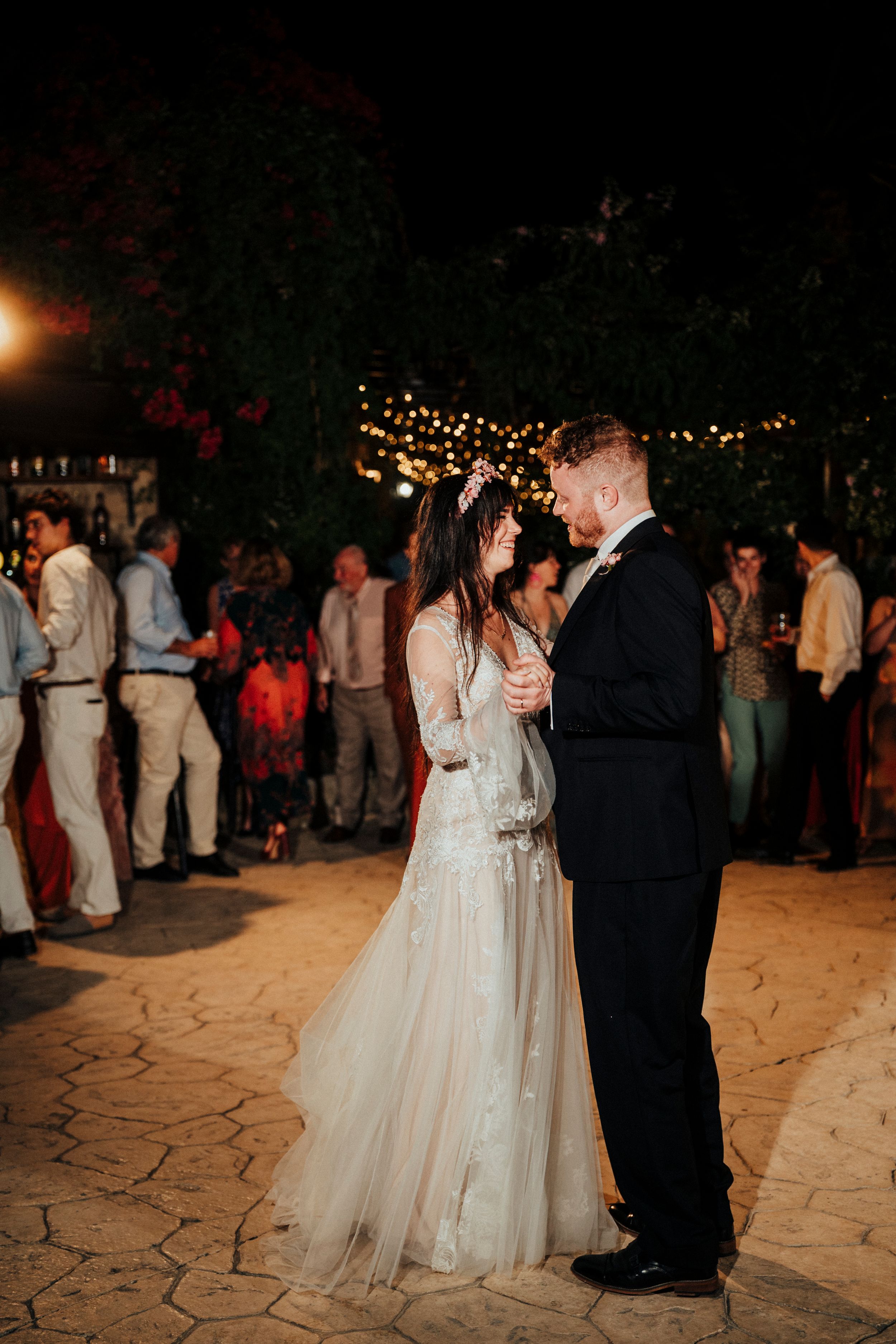 A couple shares their first dance at an outdoor evening wedding reception under twinkling lights on a stone patio.