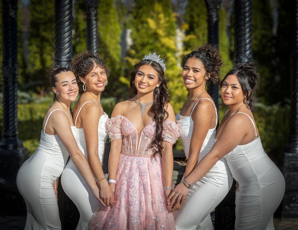Five young women pose outdoors, center girl wearing pink sparkly gown and tiara, four friends in white dresses surrounding her.