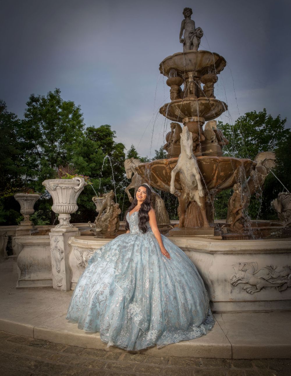 Elegant blue ballgown dress photographed in multiple angles against ornate stone fountain and garden setting at sunset.