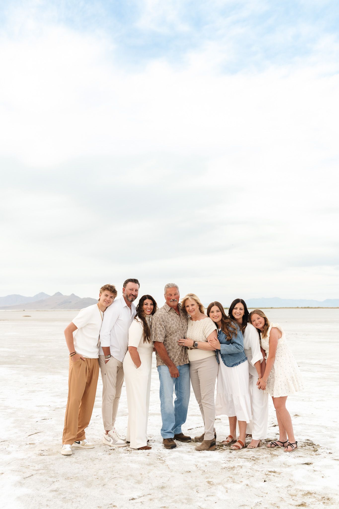 A joyful family of seven poses together on a white salt flat under a cloudy sky, dressed in coordinated neutral outfits.