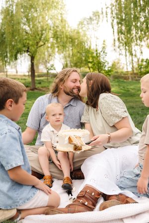 Family celebrating with a white cake outdoors on a blanket under willow trees, parents sharing a kiss while children watch.
