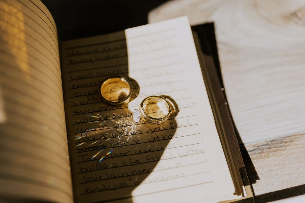 Two gold wedding bands resting on handwritten vow book pages in warm sunlight.