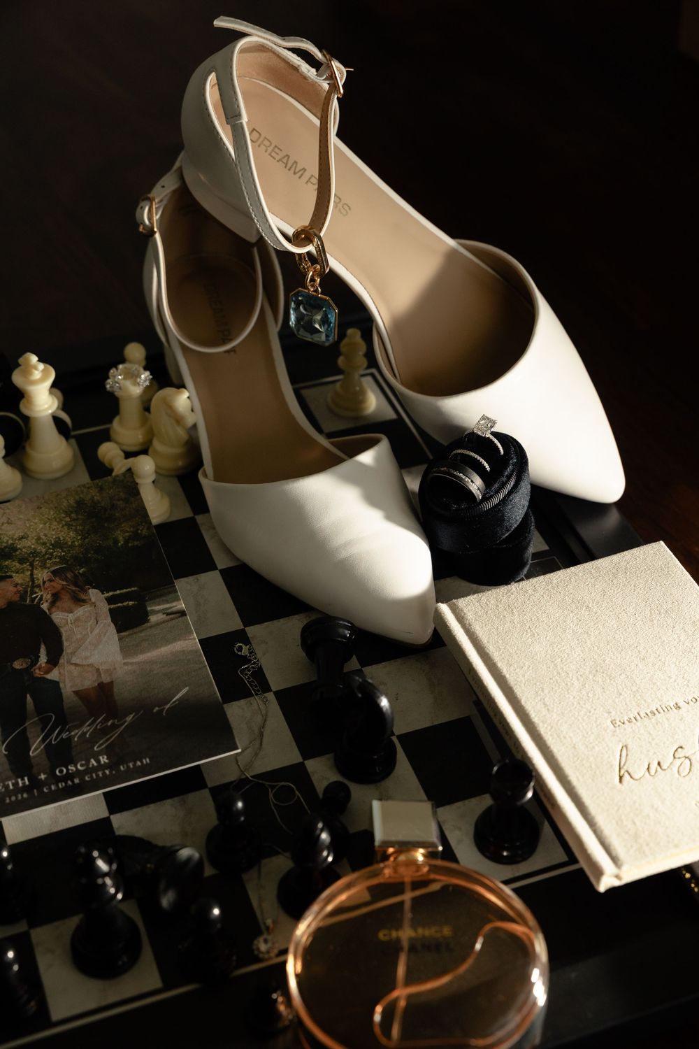 White wedding heels on chess board with Chanel perfume and vow book in moody lighting.