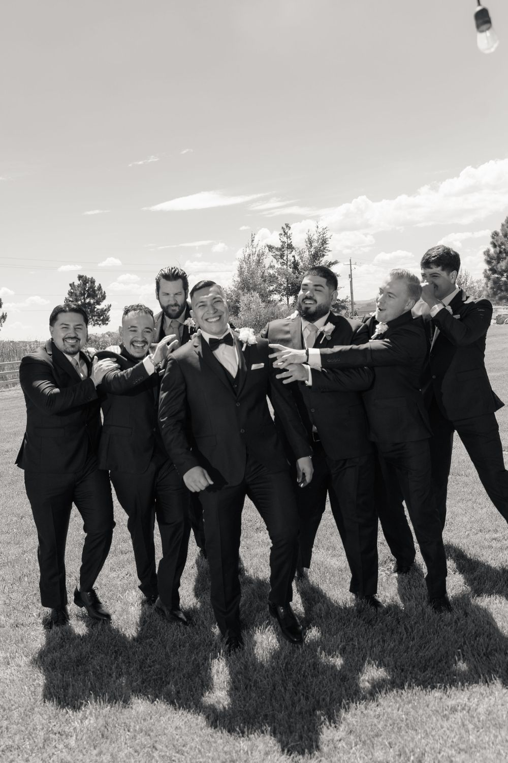 Black and white photo of groom and groomsmen in tuxedos posing playfully on a lawn.