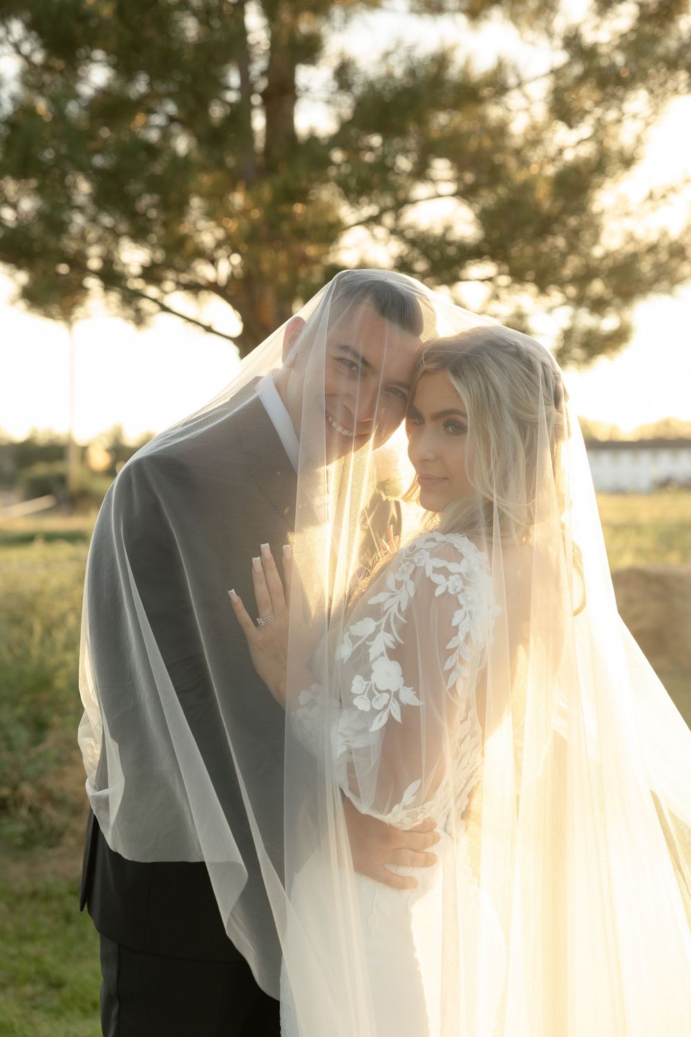 Bride and groom sharing romantic moment under flowing veil at golden sunset outdoor wedding.