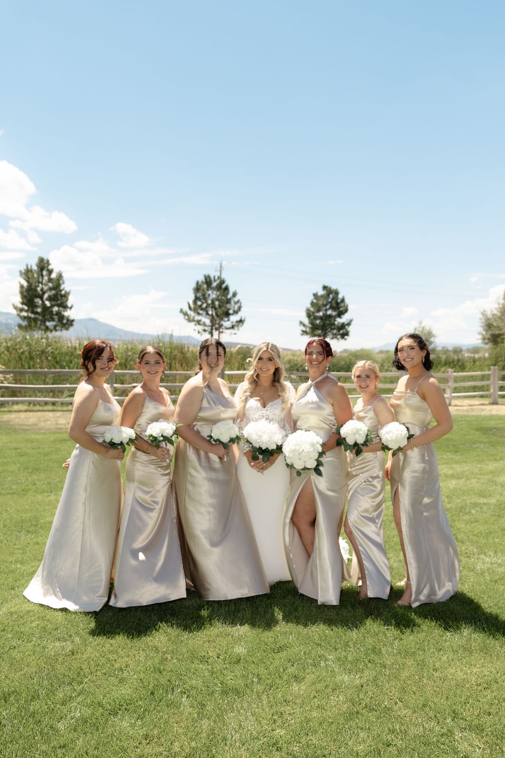 Bride with six bridesmaids in champagne gowns holding white bouquets on sunny lawn.
