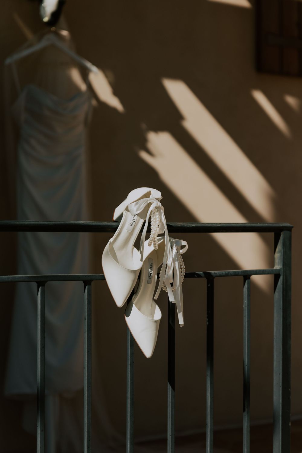 White bridal heels hanging on iron railing with wedding dress in soft background light.