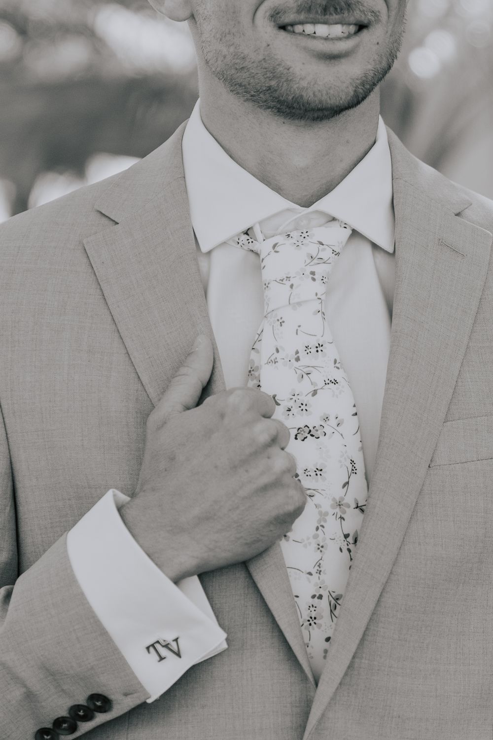 Black and white close-up of groom in suit adjusting floral tie with monogrammed cufflinks.